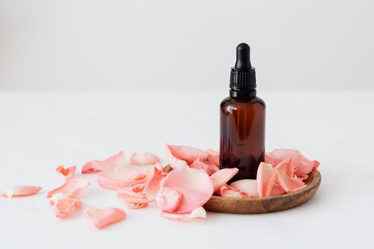 Gentle pale pink wavy rose petals placed on small round wooden plate and table near dark glass essence flask on white background